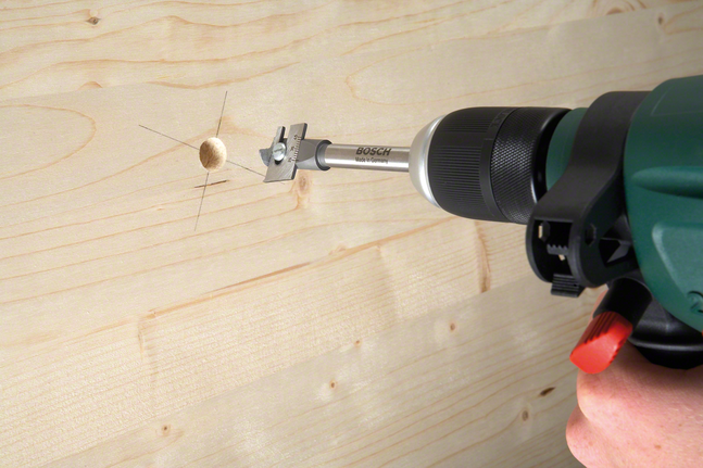 Person using a drill with a flat drill bit to bore a hole in wooden board.