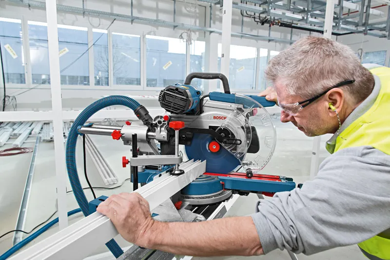 A person wearing safety equipment cuts a metal profile using a sliding mitre saw.