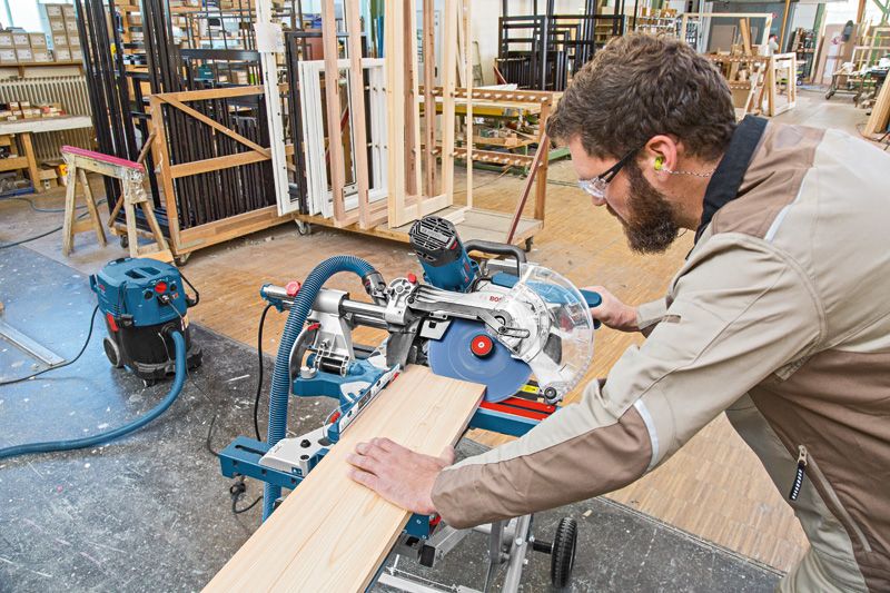 Person wearing safety equipment cuts wood using a sliding mitre saw in a workshop.