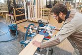 Person wearing safety equipment cuts wood using a sliding mitre saw in a workshop.
