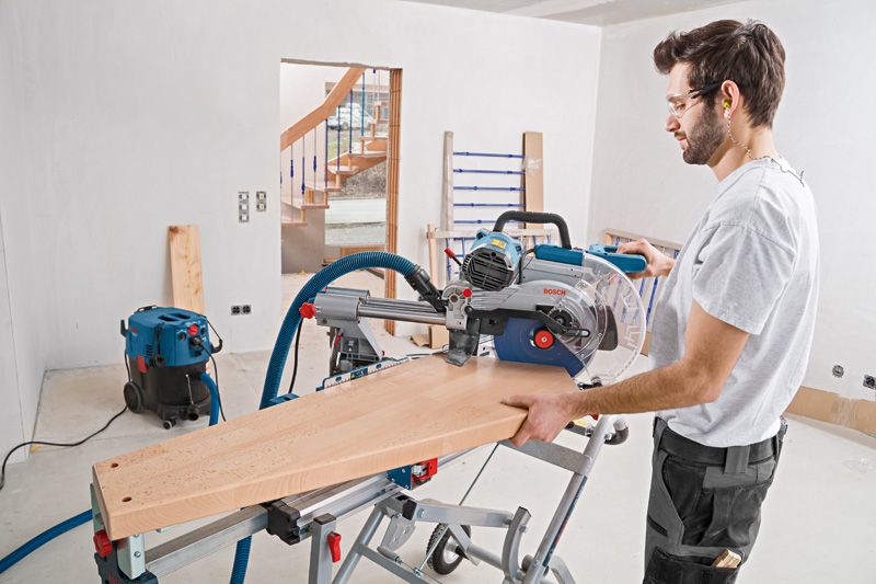Person wearing safety equipment cuts wood using a sliding mitre saw in a workshop.