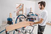 Person wearing safety equipment cuts wood using a sliding mitre saw in a workshop.