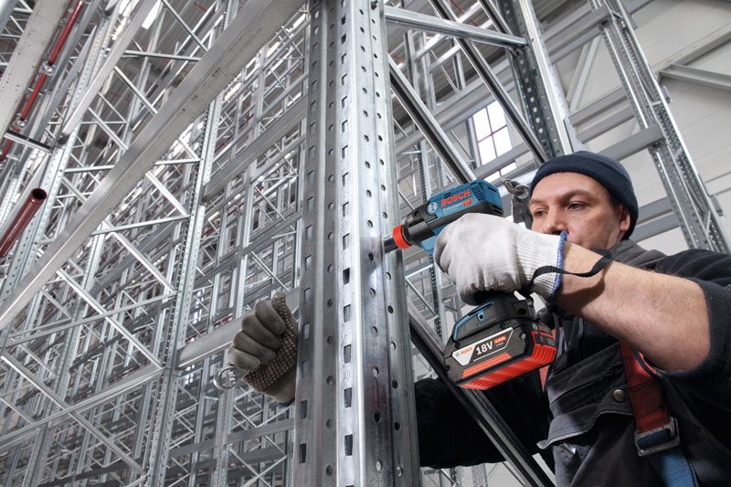 A person wearing safety equipment uses an impact driver on a metal support beam.