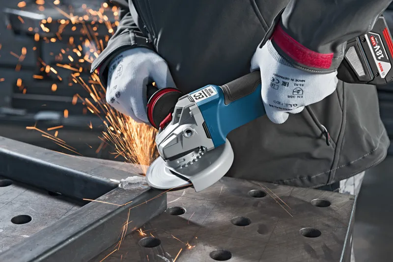 A person wearing safety equipment grinds metal with a cordless angle grinder.