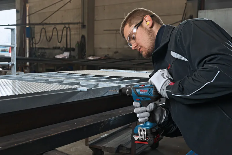 A person wearing safety equipment drills into a metal beam in an industrial setting.