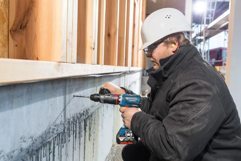 A person wearing safety equipment drills into a concrete wall at a construction site.