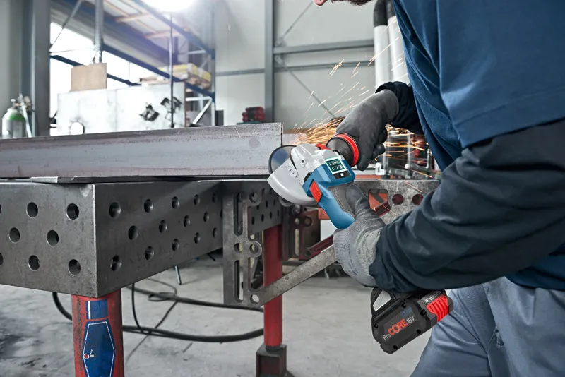 A person wearing safety equipment grinds a metal beam with a cordless angle grinder.
