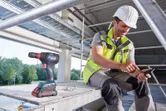 A person wearing safety equipment uses a smartphone beside a cordless drill on a construction site.