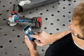 A person wearing safety equipment uses a smartphone to control a cordless angle grinder.