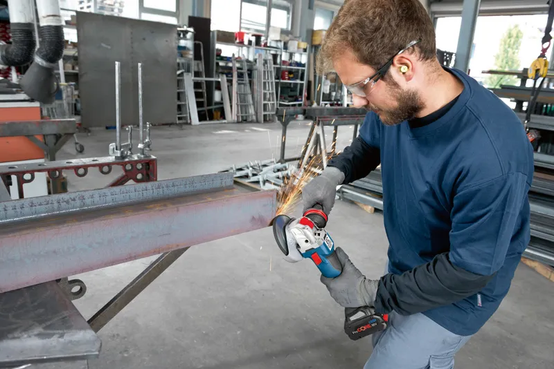 A person wearing safety equipment grinds a steel beam with a cordless angle grinder.