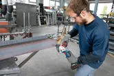 A person wearing safety equipment grinds a steel beam with a cordless angle grinder.