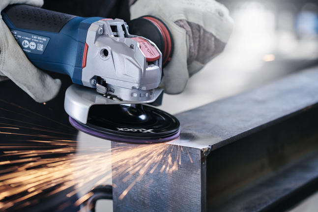 A person wearing safety equipment grinds a metal beam with an angle grinder, producing sparks.
