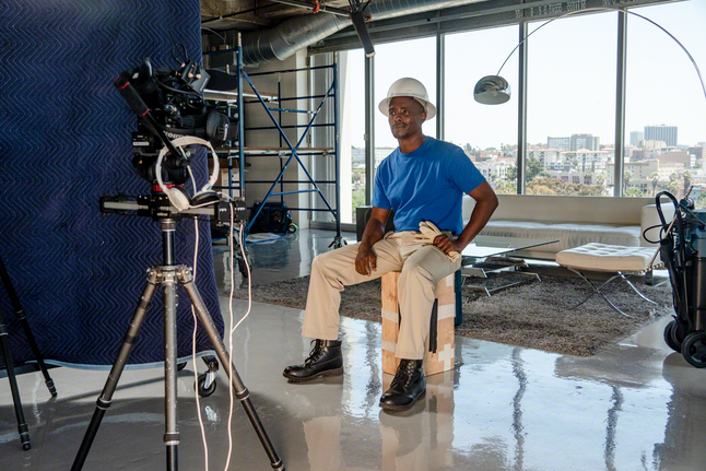 A person wearing safety equipment sits on a wooden box being filmed in a modern office.