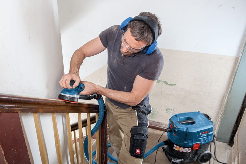 A person wearing safety equipment sands a wooden handrail with a random orbit sander.
