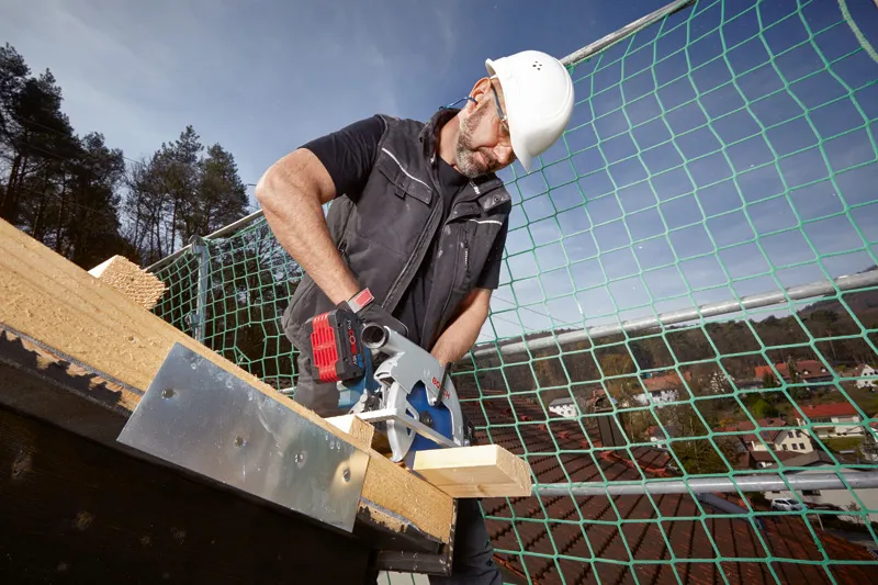 A person wearing safety equipment uses a circular saw to cut wood on a rooftop.