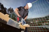 A person wearing safety equipment uses a circular saw to cut wood on a rooftop.