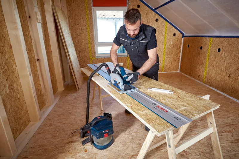A person wearing safety equipment cuts chipboard with a cordless circular saw on a guide rail.