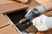 A person wearing safety equipment uses a cordless drill driver on wooden decking.