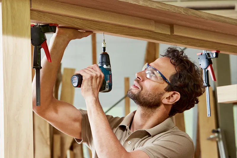 A person wearing safety equipment uses a cordless drill to fasten a screw into wooden beams.