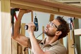 A person wearing safety equipment uses a cordless drill to fasten a screw into wooden beams.