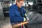 A person wearing safety equipment grinds a metal beam with an angle grinder in a workshop.
