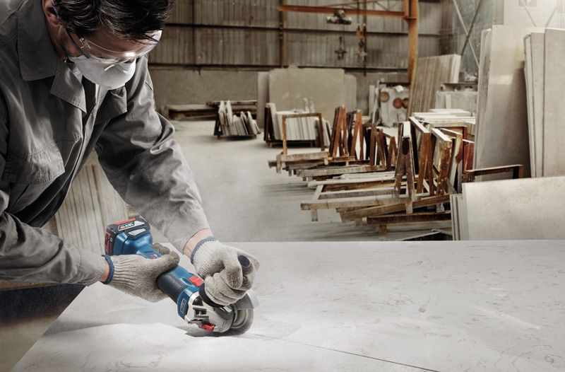 A person wearing safety equipment cuts a slab with a cordless angle grinder in a workshop.