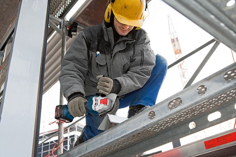 A person wearing safety equipment uses a cordless angle grinder to cut metal on scaffolding.