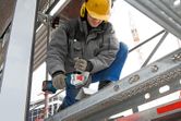A person wearing safety equipment uses a cordless angle grinder to cut metal on scaffolding.