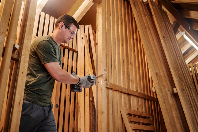 Person wearing safety equipment uses a reciprocating saw to cut wood in a workshop.