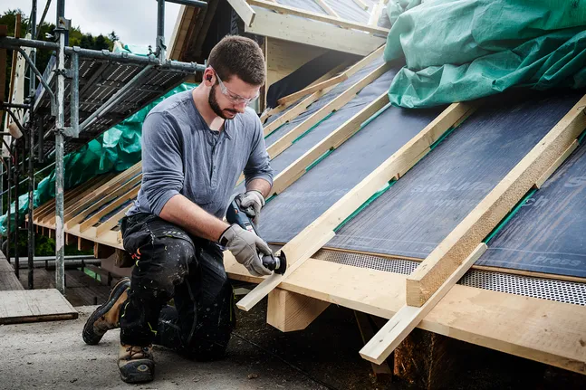 A person wearing safety equipment cuts a wooden board with a power tool on a roof.