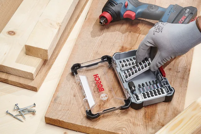Person wearing safety equipment selects a bit from a toolkit on a wooden workbench.