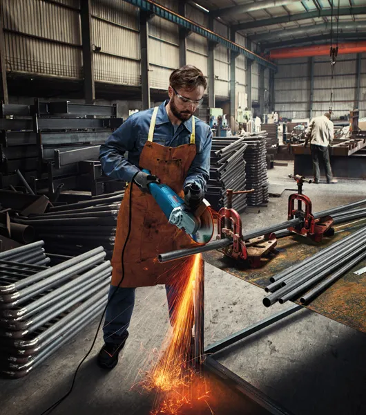 A person wearing safety equipment cuts a metal pipe with an angle grinder, sparks flying.