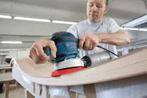 A person wearing safety equipment sands a curved wooden surface with a power tool.