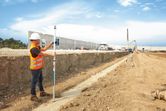 A person wearing safety equipment uses a laser receiver to measure ground level at a construction site.