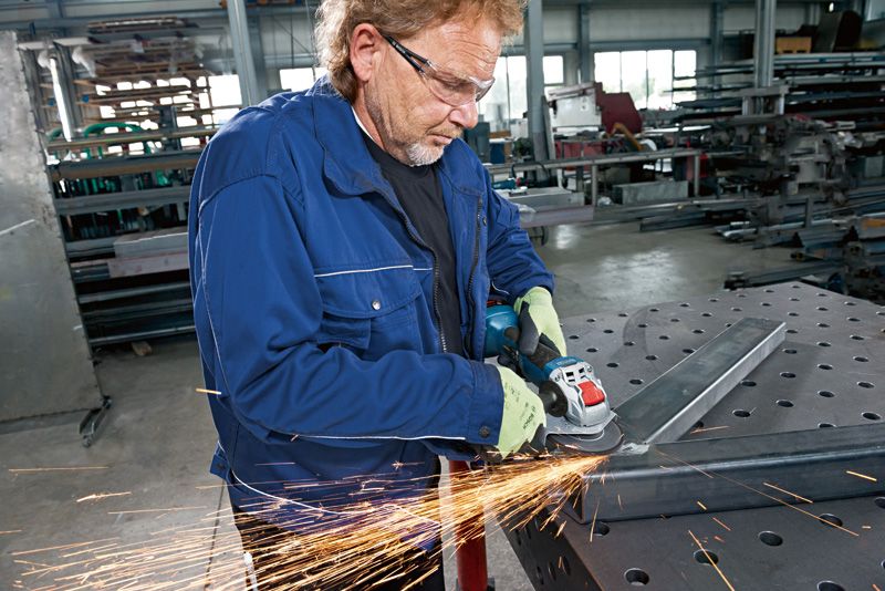 A person wearing safety equipment grinds metal with a cordless angle grinder.