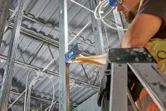 A person wearing safety equipment uses a laser receiver on a metal beam inside a building.