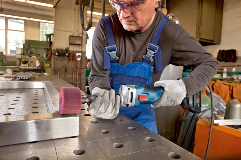 A person wearing safety equipment uses a cordless straight grinder to polish a metal edge.