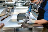 A person wearing safety equipment grinds a metal sheet with a cordless straight grinder.