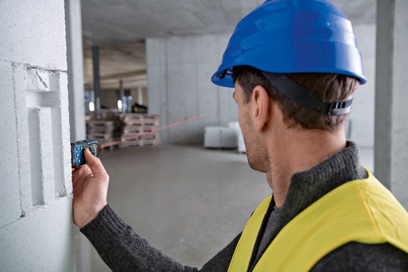 A person wearing safety equipment uses a laser measure against a concrete wall.