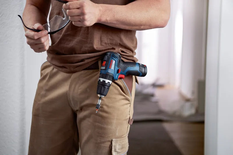 A person holding safety glasses with a cordless combi tool clipped to their pocket.