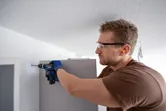 A person wearing safety equipment drills into a wall while installing furniture.