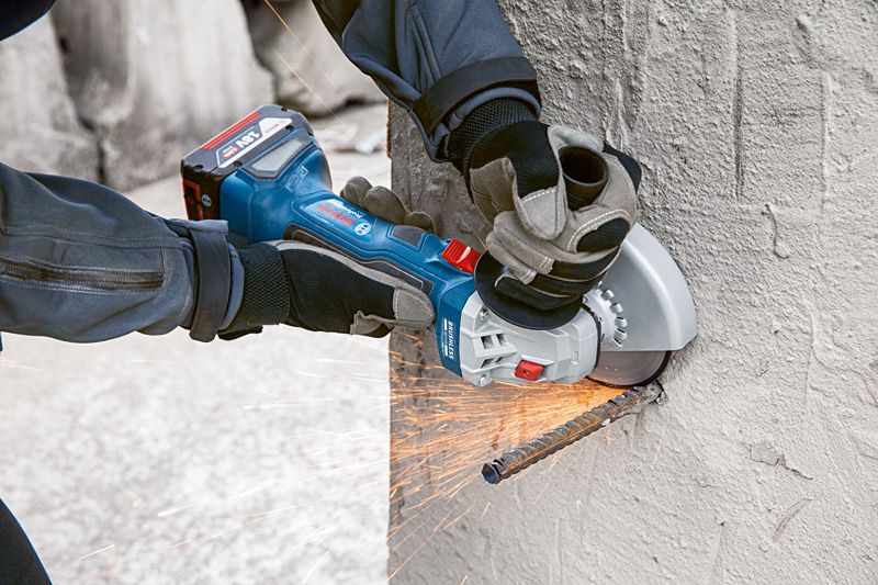A person wearing safety equipment uses an angle grinder to cut a rebar in concrete.