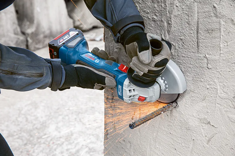 A person wearing safety equipment uses an angle grinder to cut a rebar in concrete.