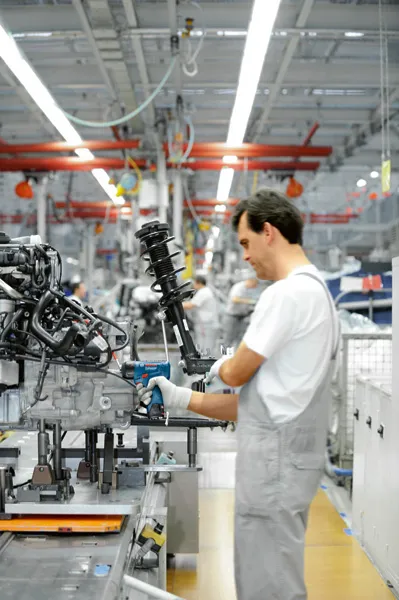 Person wearing safety equipment uses a power tool to assemble machinery on a factory line.