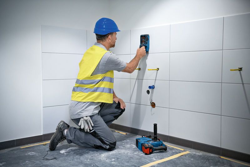 A person wearing safety equipment uses a wall scanner on tiled wall.