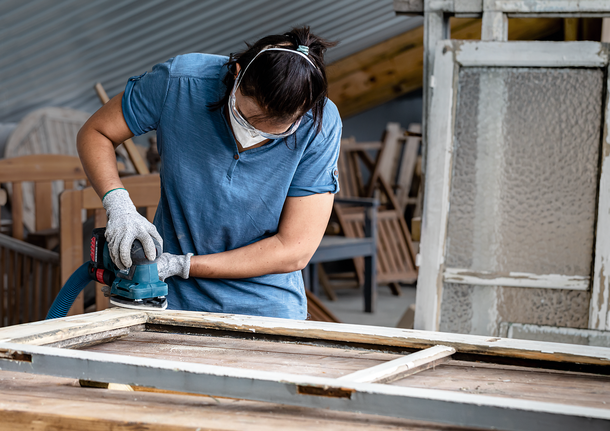 Person wearing safety equipment sands an old window frame with a power sander.