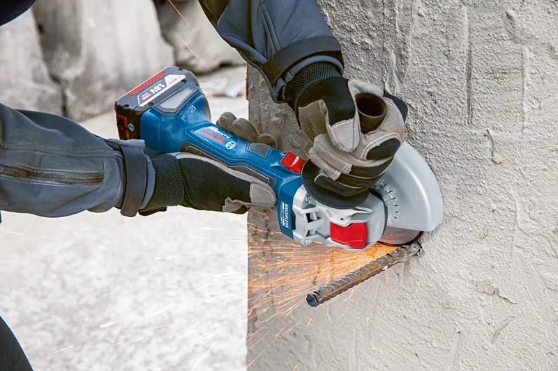 A person wearing safety equipment cuts a concrete wall with a cordless angle grinder.