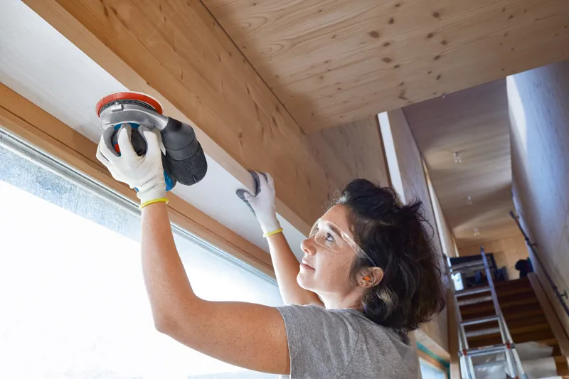 A person wearing safety equipment uses a sander to smooth a wooden beam indoors.