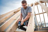 Person wearing safety equipment uses a cordless impact wrench to drive a screw into a wooden frame.