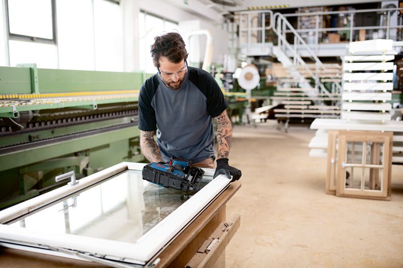 A person wearing safety equipment uses a cordless wood nailer to secure a window frame.